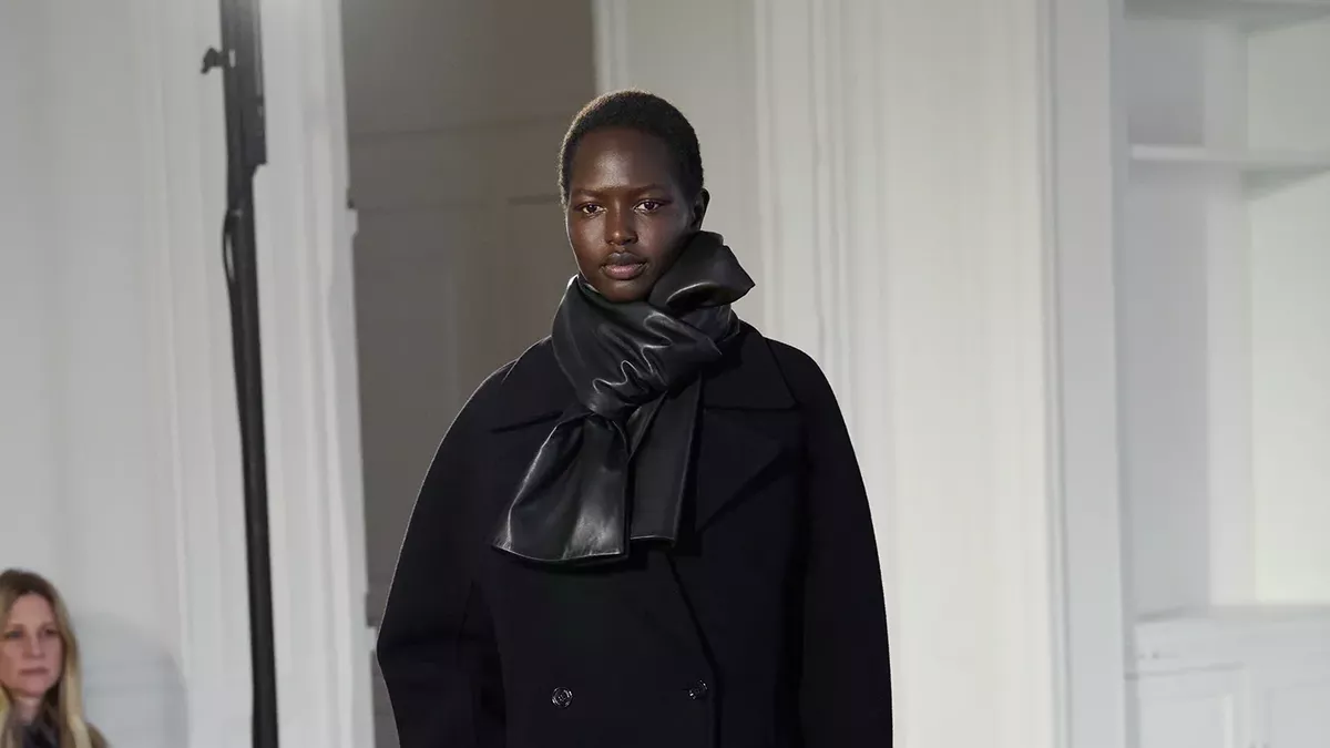 Model walking a fashion runway in a black tailored double-breasted coat paired with a black leather scarf.