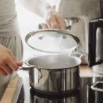 Young man cooking fresh food at home and opening lid of steaming pot