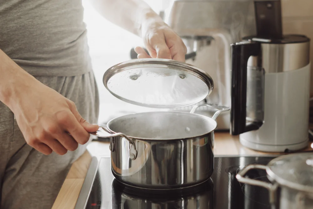 Young man cooking fresh food at home and opening lid of steaming pot