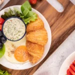 Breakfast consists of croissant, fried egg, salad dressing, black grapes, and tomatoes.