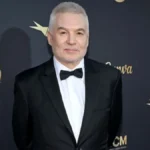 man in a tuxedo stands smiling against a dark backdrop