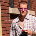 Justin Bieber Net Worth 2026 showing Justin Bieber standing outdoors wearing sunglasses and a checkered shirt near a brick wall.