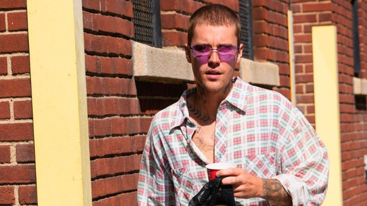 Justin Bieber Net Worth 2026 showing Justin Bieber standing outdoors wearing sunglasses and a checkered shirt near a brick wall.