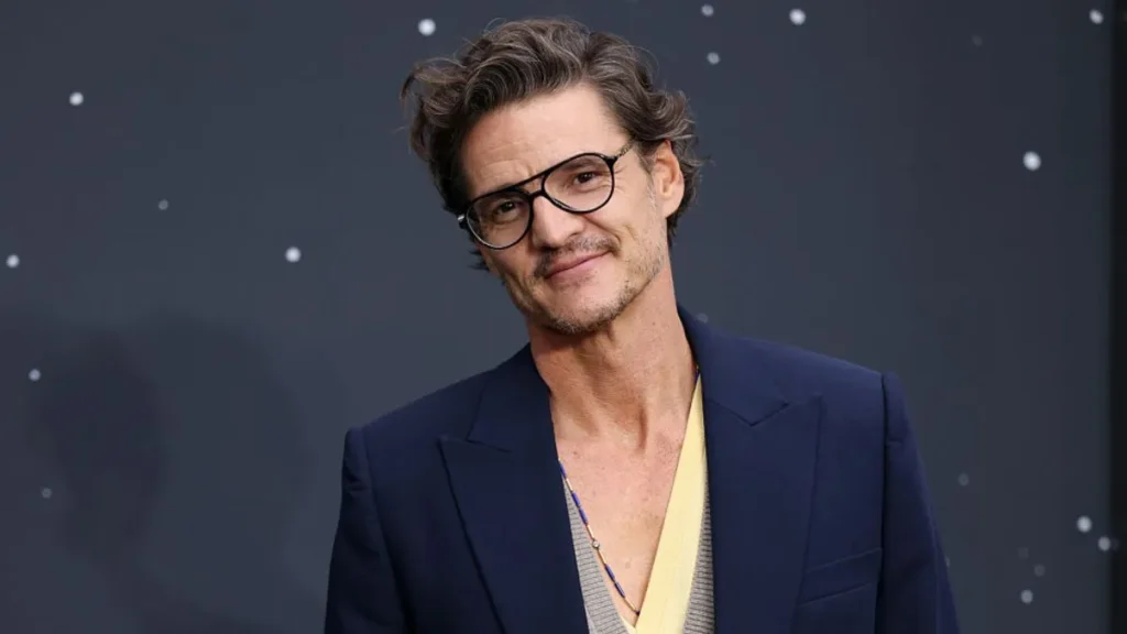 Pedro Pascal wearing glasses and a navy blazer at a red-carpet event, smiling at the camera.