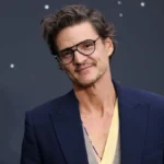 Pedro Pascal wearing glasses and a navy blazer at a red-carpet event, smiling at the camera.