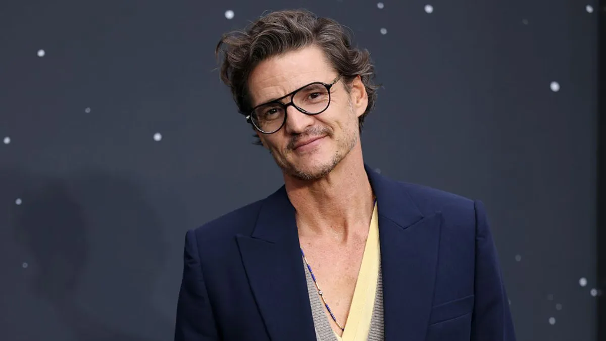 Pedro Pascal wearing glasses and a navy blazer at a red-carpet event, smiling at the camera.