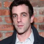 B.J. Novak Net Worth featured in a portrait of the actor and writer wearing a dark coat and button-up shirt standing in front of a red brick wall background.