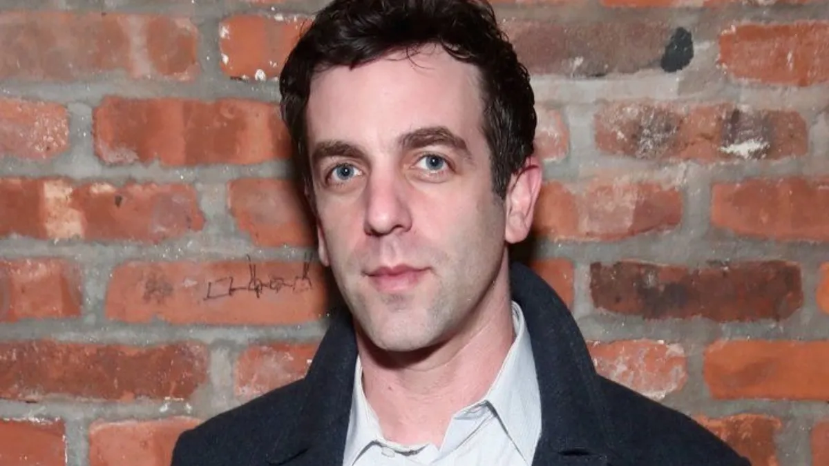 B.J. Novak Net Worth featured in a portrait of the actor and writer wearing a dark coat and button-up shirt standing in front of a red brick wall background.
