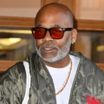 dame dash net worth highlighted in a portrait of Damon Dash wearing orange-tinted sunglasses, a white shirt, and an olive green quilted jacket.