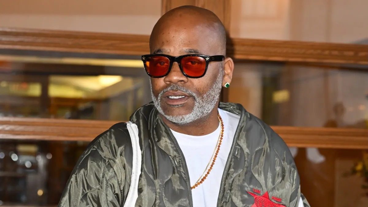 dame dash net worth highlighted in a portrait of Damon Dash wearing orange-tinted sunglasses, a white shirt, and an olive green quilted jacket.