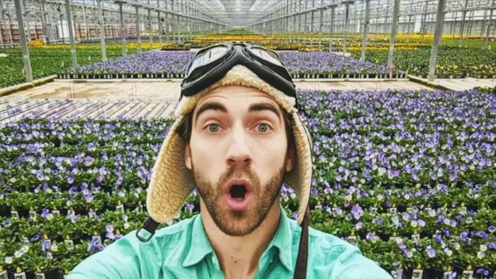 Danny Go Net Worth featured in a selfie of children's host Danny Go making a surprised face inside a massive greenhouse surrounded by purple flowers.