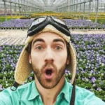 Danny Go Net Worth featured in a selfie of children's host Danny Go making a surprised face inside a massive greenhouse surrounded by purple flowers.
