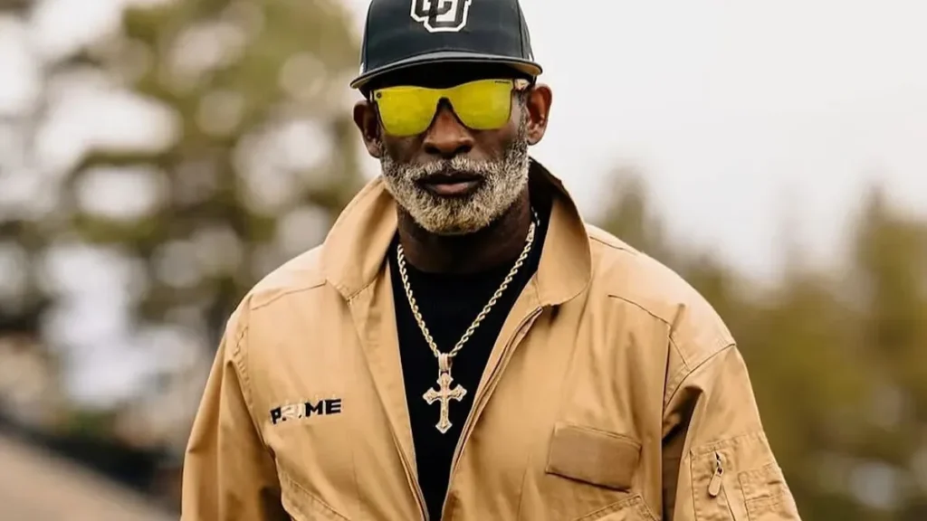 Deion Sanders wearing Coach Prime gear and yellow sunglasses outdoors.featuring Deion Sanders net worth