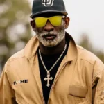 Deion Sanders wearing Coach Prime gear and yellow sunglasses outdoors.featuring Deion Sanders net worth