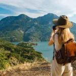 A female traveler wearing a hat and backpack taking photos of a scenic lake surrounded by lush green mountains.