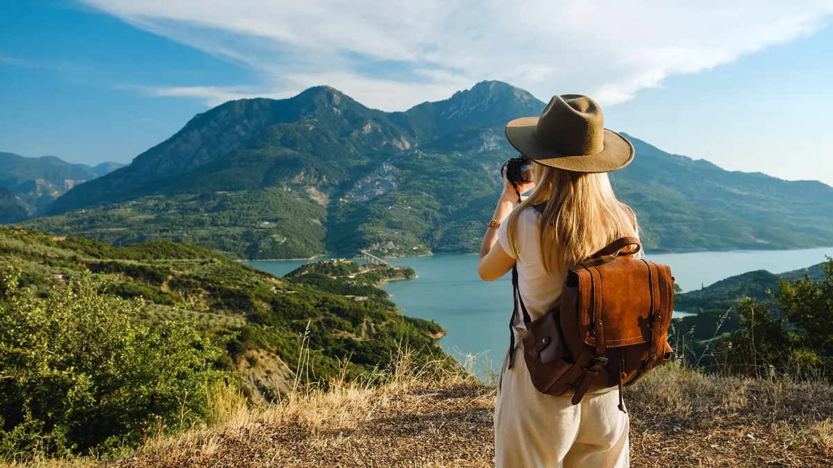 A female traveler wearing a hat and backpack taking photos of a scenic lake surrounded by lush green mountains.