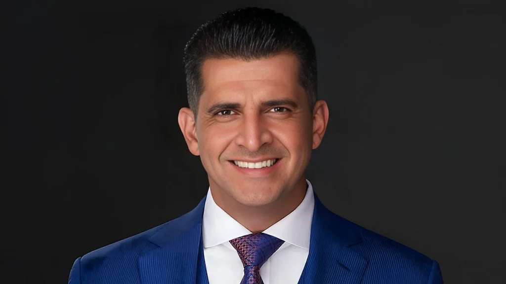 A headshot of a smiling, dark-haired man in a blue suit jacket, white dress shirt, and patterned blue tie set against a dark background.