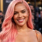 Karol g net worth highlighted in a portrait of Karol G with long pink hair smiling in a burnt orange satin gown at a red carpet event.