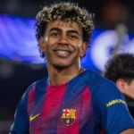 A close-up portrait of FC Barcelona footballer Lamine Yamal wearing his blue and red home kit, smiling with curly highlighted hair and braces, ideal for an article discussing lamine yamal net worth.