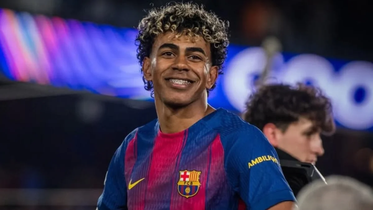 A close-up portrait of FC Barcelona footballer Lamine Yamal wearing his blue and red home kit, smiling with curly highlighted hair and braces, ideal for an article discussing lamine yamal net worth.