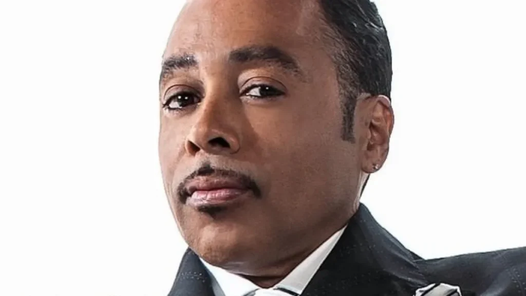 Morris Day Net Worth featured in a headshot portrait of the singer wearing a black pinstripe suit with a striped tie and matching pocket square.