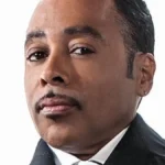 Morris Day Net Worth featured in a headshot portrait of the singer wearing a black pinstripe suit with a striped tie and matching pocket square.